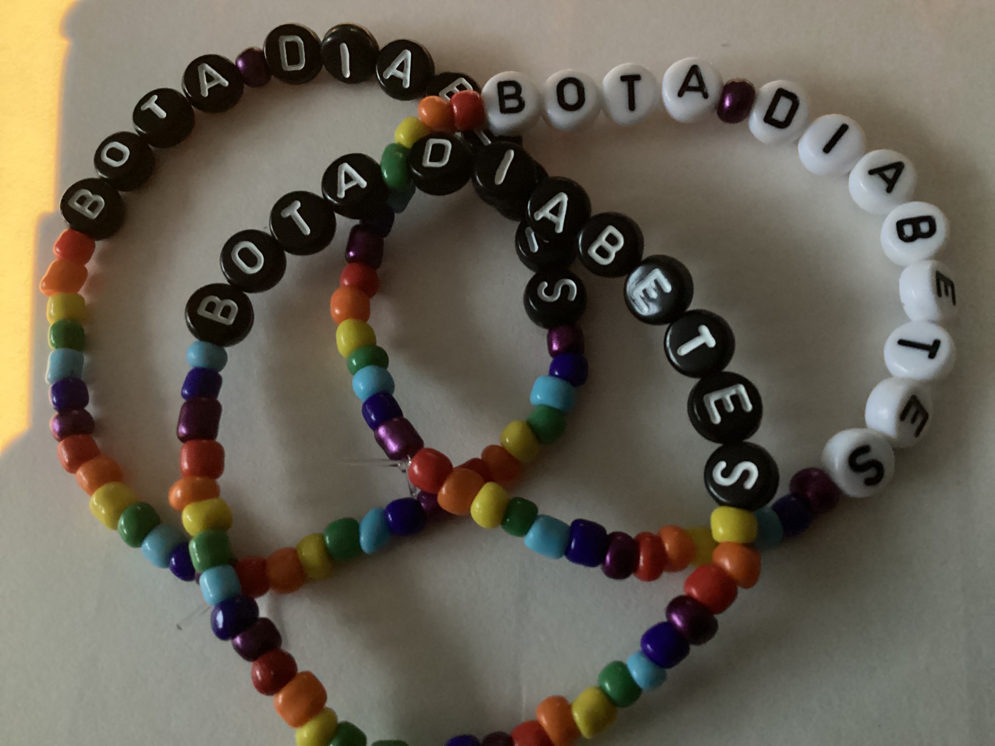 Tre elastiska budskapsarmband med bokstavspärlor och regnbågsfärgade småpärlor som bildar text.
