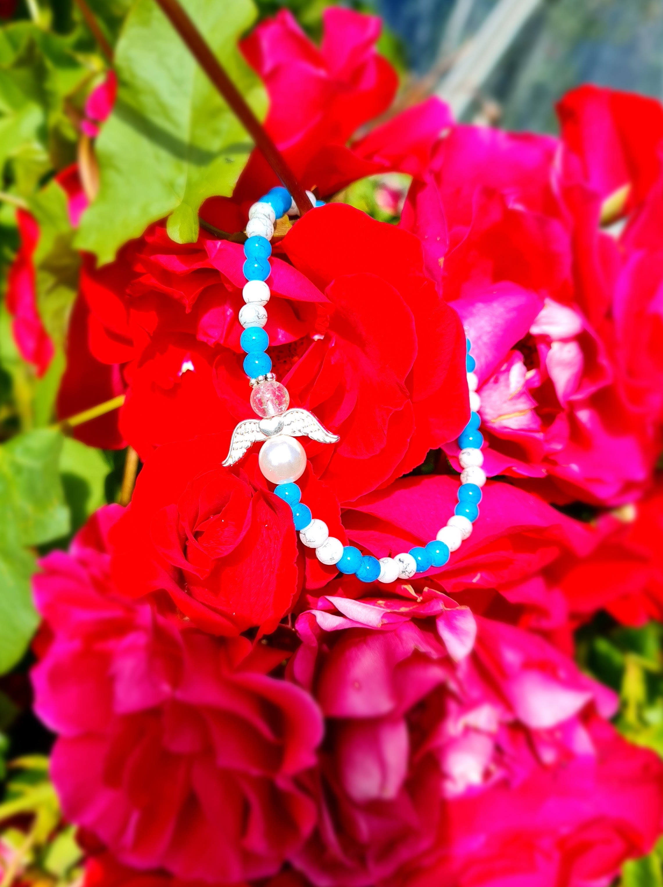 Ängelarmband i blått och vitt med vingar och mittpärla, fotat mot röda blommor.